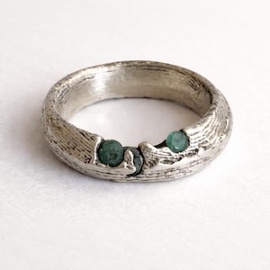 Könnte beinhalten: Ein silberner Ring mit strukturierter Oberfläche und drei grünen Edelsteinen. Der Ring hat ein rustikales, organisches Design, wobei die Edelsteine in das Band eingesetzt sind. Der Ring ist ein Schmuckstück.