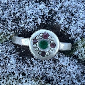 Peut inclure: Une bague en argent avec un motif circulaire. Le centre présente une pierre verte, entourée d'un anneau de petites perles rondes et de quatre pierres marron. La bague est posée sur un fond givré texturé.