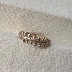Könnte beinhalten: Ein goldfarbener Ring mit einem einzigartigen Design. Der Ring weist eine Reihe von vertikalen Stäben auf, die durch horizontale Stäbe verbunden sind und ein strukturiertes, geometrisches Muster bilden. Der Ring ist vor einem weißen Hintergrund platziert.