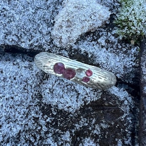 Könnte beinhalten: Ein silberner Ring mit strukturierter Oberfläche, verziert mit drei rosa Edelsteinen und einem klaren Edelstein. Der Ring liegt auf einem Hintergrund aus Eiskristallen und dunklem Stein.