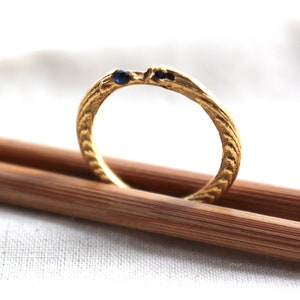 Könnte beinhalten: Ein goldener Ring mit einem strukturierten Design und zwei blauen Edelsteinen. Der Ring liegt auf zwei Holzstäbchen.