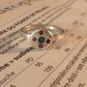 Puede incluir: Un anillo de plata con una cara circular. La cara está adornada con una piedra verde central, rodeada de pequeñas piedras rojas y un borde de diminutas cuentas plateadas. El anillo descansa sobre un fondo de papel impreso.