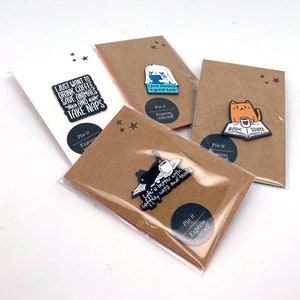 May include: Four brown card stock backing cards with black and white enamel pins featuring book and coffee themes. The pins say "I just finished a great book", "Life is better with coffee, cats and books", "Book Coffee", and "I just want to drink coffee, save animals and take naps".