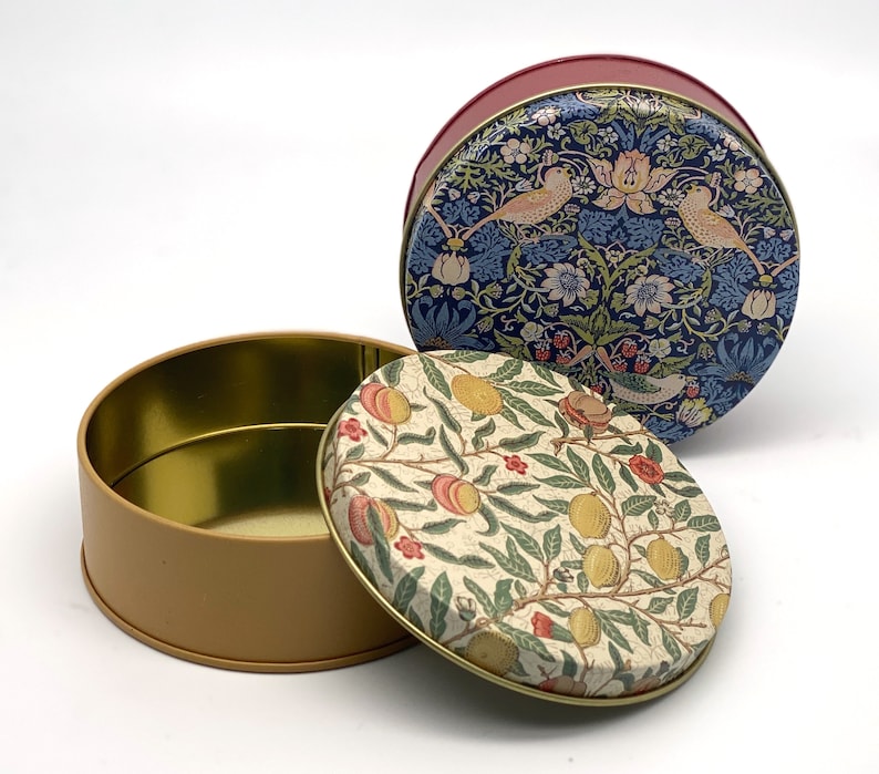 May include: Two round tin containers with floral patterns. The top container has a blue and white floral pattern with birds. The bottom container has a cream and yellow floral pattern with lemons and peaches.