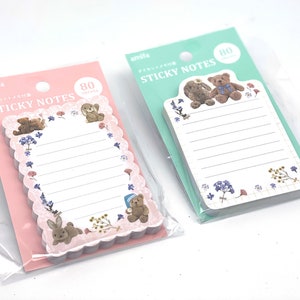 Peut inclure: Deux paquets de notes adhésives de la marque Amifa, contenant chacun 80 feuilles. Un paquet a un support rose avec un bloc-notes à bords festonnés orné d'oursons et de lapins. L'autre a un support vert menthe avec un bloc-notes rectangulaire.