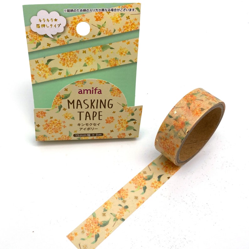 Peut inclure: Un rouleau de ruban adh&eacute;sif washi avec un motif floral dans des tons d'orange et de jaune. Le ruban mesure 15 mm de large et 3 m&egrave;tres de long. L'emballage pr&eacute;sente un fond vert avec le texte "amifa Masking Tape" et "キンモクセイ アイボリー" en japonais.