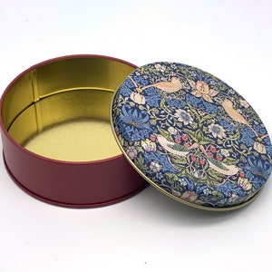 May include: A round, red tin with a gold interior and a removable lid. The lid features a floral pattern with birds in shades of blue, pink, and green.