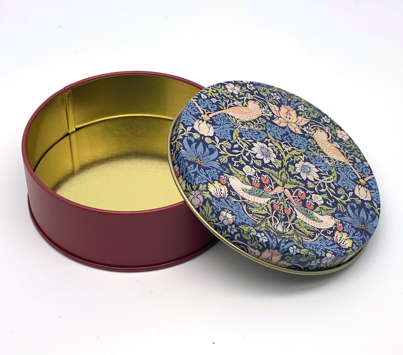 May include: A round, red tin with a gold interior and a removable lid. The lid features a floral pattern with birds in shades of blue, pink, and green.