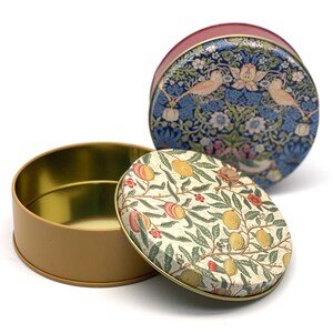 May include: Two round tin boxes with floral patterns. The top box has a blue and white floral pattern with birds. The bottom box has a cream and red floral pattern with fruit.
