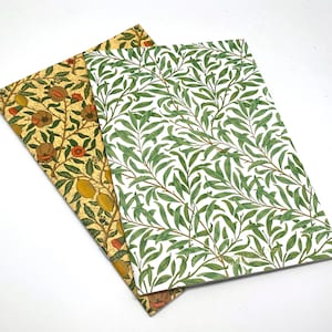 Può includere: Due quaderni con motivi floreali. Un quaderno ha uno sfondo giallo con fiori arancioni e rossi e foglie verdi. L'altro quaderno ha uno sfondo bianco con foglie verdi. Entrambi i quaderni hanno un design in foglia d'oro.