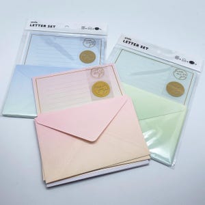 Pode incluir: Conjuntos de cartas em cores pastel, incluindo rosa, azul e verde, com envelopes a condizer. Cada conjunto é embalado numa manga de plástico transparente e inclui papel de carta pautado e um selo dourado. O texto "amila LETTER SET" está impresso na embalagem.