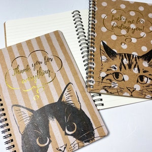 Puede incluir: Dos cuadernos de espiral con cubiertas marrones y detalles en dorado. Las cubiertas presentan ilustraciones en blanco y negro de gatos con globos de pensamiento que dicen "Gracias por todo" y "¡Qué suerte la mía por encontrarte!"