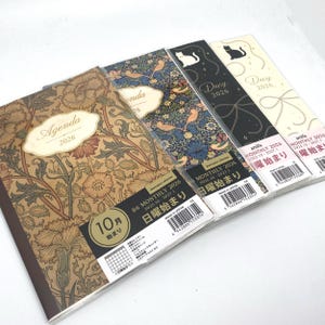 May include: A selection of 2026 agenda and diary books. The agenda has a brown floral pattern, and the diaries feature designs with birds, cats, and abstract motifs. Each book includes a label with Japanese text and the dates 2025.10-2027.1.