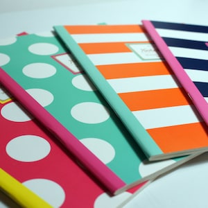 Pode incluir: Quatro cadernos com capas estampadas diferentes. O caderno de cima tem uma capa listrada azul marinho e branca com uma borda rosa. O segundo caderno tem uma capa listrada laranja e branca com uma borda verde claro. O terceiro caderno tem uma capa com bolinhas azul esverdeado e brancas com uma borda rosa. O caderno de baixo tem uma capa com bolinhas rosa e brancas com uma borda amarela.
