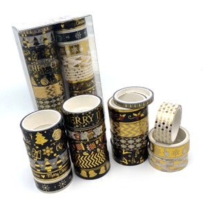 Op de afbeelding: Een verzameling decoratieve washi tapes, deels in een transparante verpakking, deels los. De tapes hebben zwarte en gouden ontwerpen met vakantiethema's, waaronder bomen, sneeuwvlokken en de tekst "Merry Christmas".