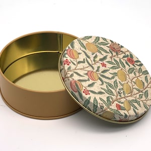 May include: A round, beige tin with a floral pattern lid. The lid features a white background with a repeating pattern of green leaves, pink flowers, and yellow fruits. The tin is empty.