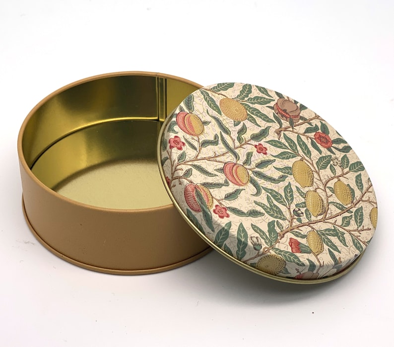 May include: A round, beige tin with a floral pattern lid. The lid features a white background with a repeating pattern of green leaves, pink flowers, and yellow fruits. The tin is empty.