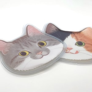 May include: Two cat-shaped gel packs. One is grey with yellow eyes, the other is calico with green eyes. Both have a clear plastic border and are designed to resemble a cat's face. The gel packs are on a white surface.