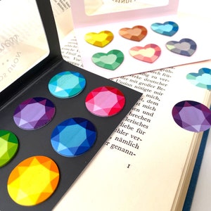 Op de afbeelding: Een set van negen kleurrijke edelsteen-vormige stickers in een regenboogkleurenschema. De stickers zijn gerangschikt in een cirkel op een zwarte achtergrond. De stickers zijn perfect om een ​​toets van glans toe te voegen aan elk project.