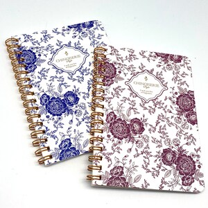 Puede incluir: Dos cuadernos de espiral con diseños florales. El cuaderno de la izquierda tiene un patrón floral azul y el cuaderno de la derecha tiene un patrón floral morado. Ambos cuadernos tienen el texto "Chinoiserie Style" y "Te deseo mucha felicidad" en la portada.