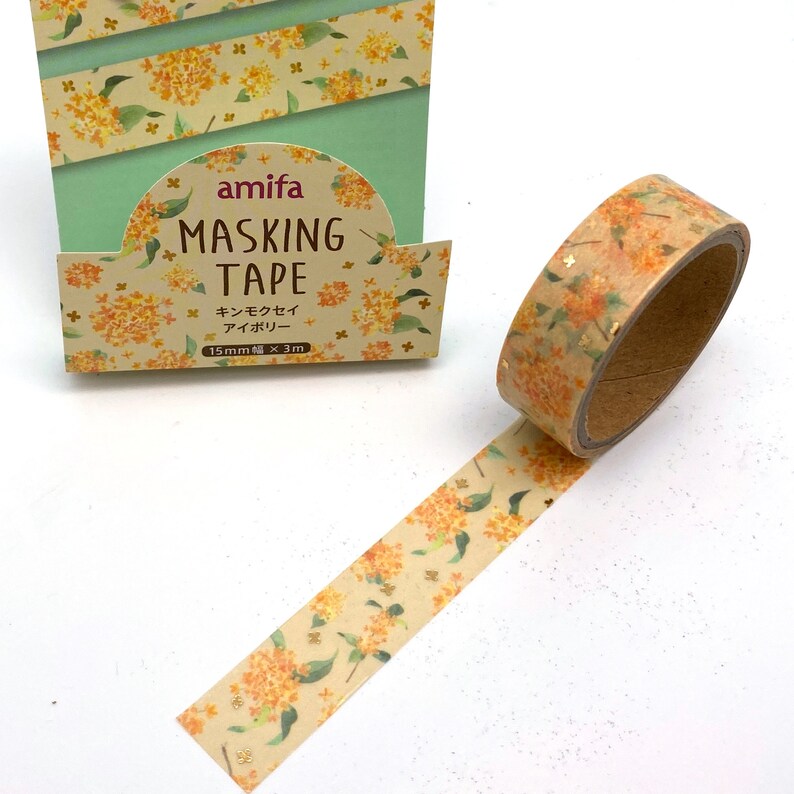 Peut inclure: Un rouleau de ruban adh&eacute;sif washi avec un motif floral. Le ruban est beige avec des fleurs orange et des paillettes dor&eacute;es. Le ruban est &eacute;tiquet&eacute; "amifa Masking Tape" et "キンモクセイ アロマリー" en japonais. Le ruban mesure 15 mm de large et 3 m de long.