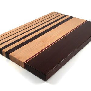 May include: Rectangular wooden cutting board with a light wood grain and dark brown stripes. The board has a section of solid dark brown wood with a thin red stripe. A kitchen accessory.