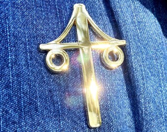 May include: A gold-colored metal pin in the shape of a stylized cross with two circles on either side of the cross. The pin is worn on a blue denim shirt.