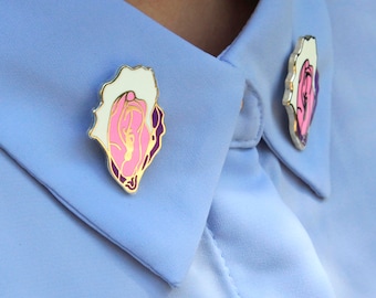 May include: Two gold-colored pins with pink and white enamel designs are attached to a light blue collared shirt.