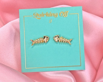 May include: Gold-colored fish skeleton earrings displayed on a teal card with the text "Stalking Off". The card is set against a pink satin background. The earrings are a unique accessory.