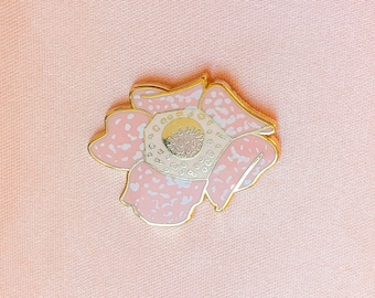 May include: A gold-tone enamel pin featuring a pink flower with white speckles. The center of the flower has a gold-tone circle with a white design.
