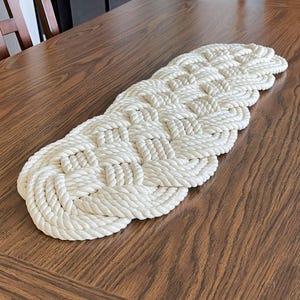 May include: A white rope table runner with a woven knot design. The runner is on a wooden table.