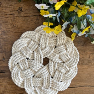 May include: A white, round, woven rope coaster with a knot design. The coaster is made of thick, natural rope and has a rustic, handmade look.