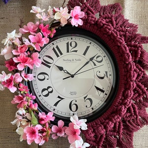 May include: A burgundy macrame wall hanging with a black clock face featuring the text "Sterling & Noble Clock Company". The clock is surrounded by a wreath of pink and white artificial flowers.