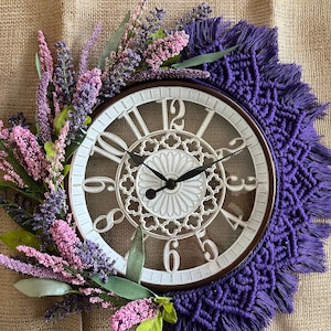May include: A white clock with roman numerals and a decorative design is surrounded by a purple macrame wreath with fringe. The wreath is decorated with a cluster of pink and purple flowers.