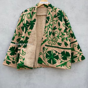 Op de afbeelding: Een beige en groene, met bloemen geborduurde jas met een sherpa-voering. De jas heeft een kimono-stijl met wijde mouwen en een sluiting aan de voorkant.