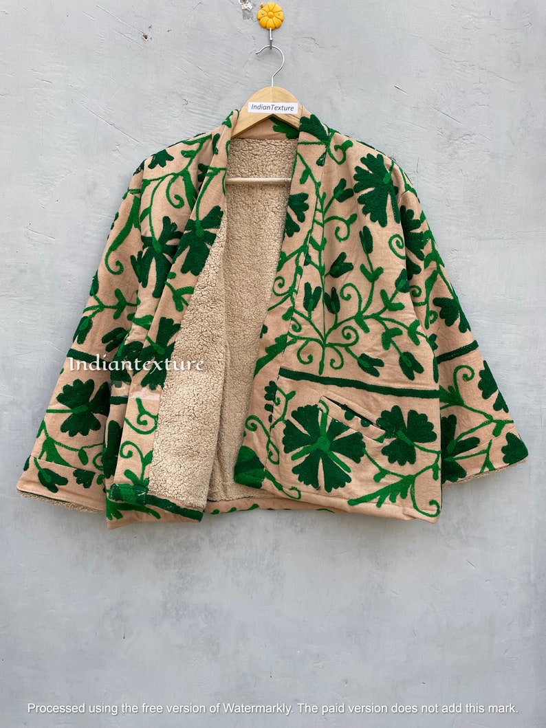 Op de afbeelding: Een beige en groene, met bloemen geborduurde jas met een sherpa-voering. De jas heeft een kimono-stijl met wijde mouwen en een sluiting aan de voorkant.