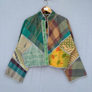 Peut inclure: Une veste courte présentant un motif patchwork composé de divers tissus dans des tons de vert, jaune, marron et crème. Le vêtement possède des manches longues, un col officier et des coutures visibles, avec un liseré gris clair sur les bords.