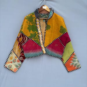 Op de afbeelding: Een kort jasje gemaakt van patchworkstof met een verscheidenheid aan bloemmotieven en geometrische patronen in levendige tinten geel, groen, roze, oranje en wit. Het kledingstuk heeft lange mouwen, een opstaande kraag en een zwart-witte bies.