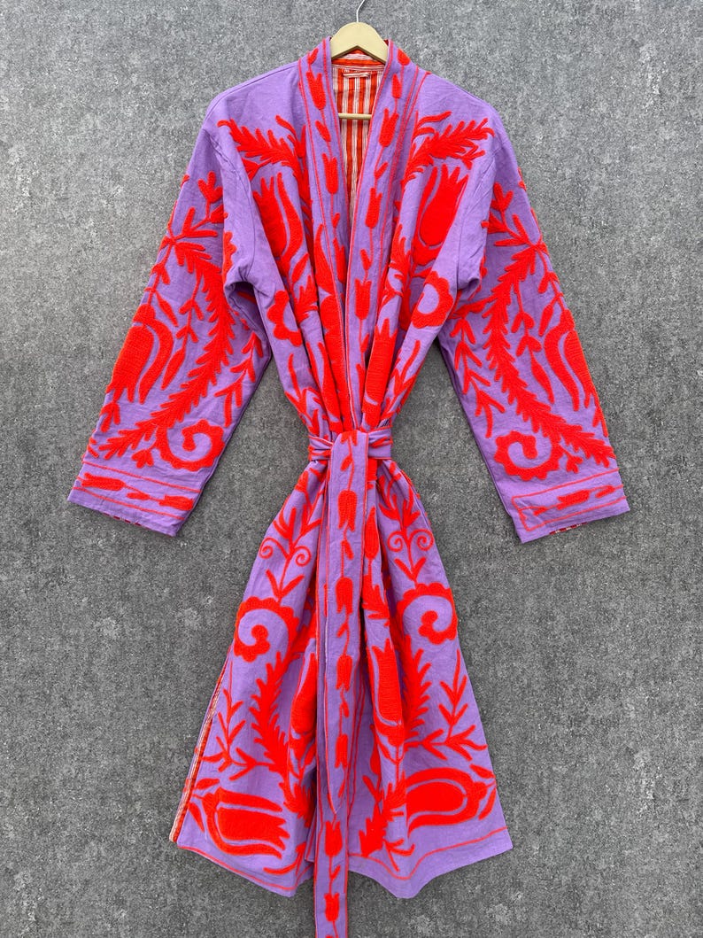 Puede incluir: Una bata morada con un estampado floral rojo. La bata tiene un cintur&oacute;n de atar y un forro a rayas rojo y blanco.