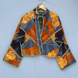 Könnte beinhalten: Eine kurz geschnittene Jacke mit einem Patchwork-Design aus orangefarbenen, gelben und dunkelblauen Stoffbahnen mit Blattmustern und geometrischen Formen. Das Kleidungsstück hat lange Ärmel, eine offene Vorderseite und hellgrüne Kanten.