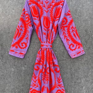 Puede incluir: Una bata morada con un patr&oacute;n de bordado floral rojo. La bata tiene mangas largas y un cintur&oacute;n de atar.