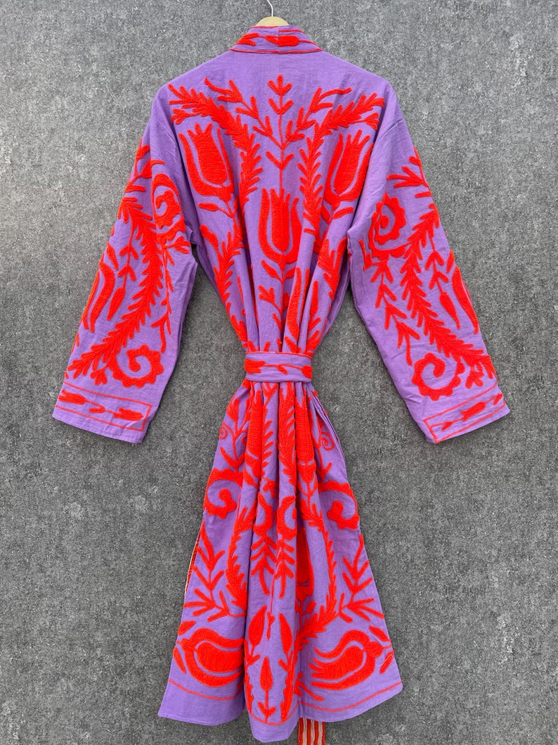 Puede incluir: Una bata morada con un patr&oacute;n de bordado floral rojo. La bata tiene mangas largas y un cintur&oacute;n de atar.