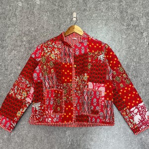 Op de afbeelding: Een jas met lange mouwen en een patchworkontwerp in tinten rood, oranje en wit. Het kledingstuk toont diverse bloem- en geometrische patronen over de panelen, een knoopsluiting aan de voorzijde en een standaard kraag op een kledinghanger.