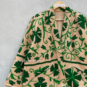 Op de afbeelding: Een beige jas met een groen geborduurd bloemenpatroon. De jas heeft een shawlkraag en lange mouwen. De jas is gevoerd met een zacht, bruin fleece.