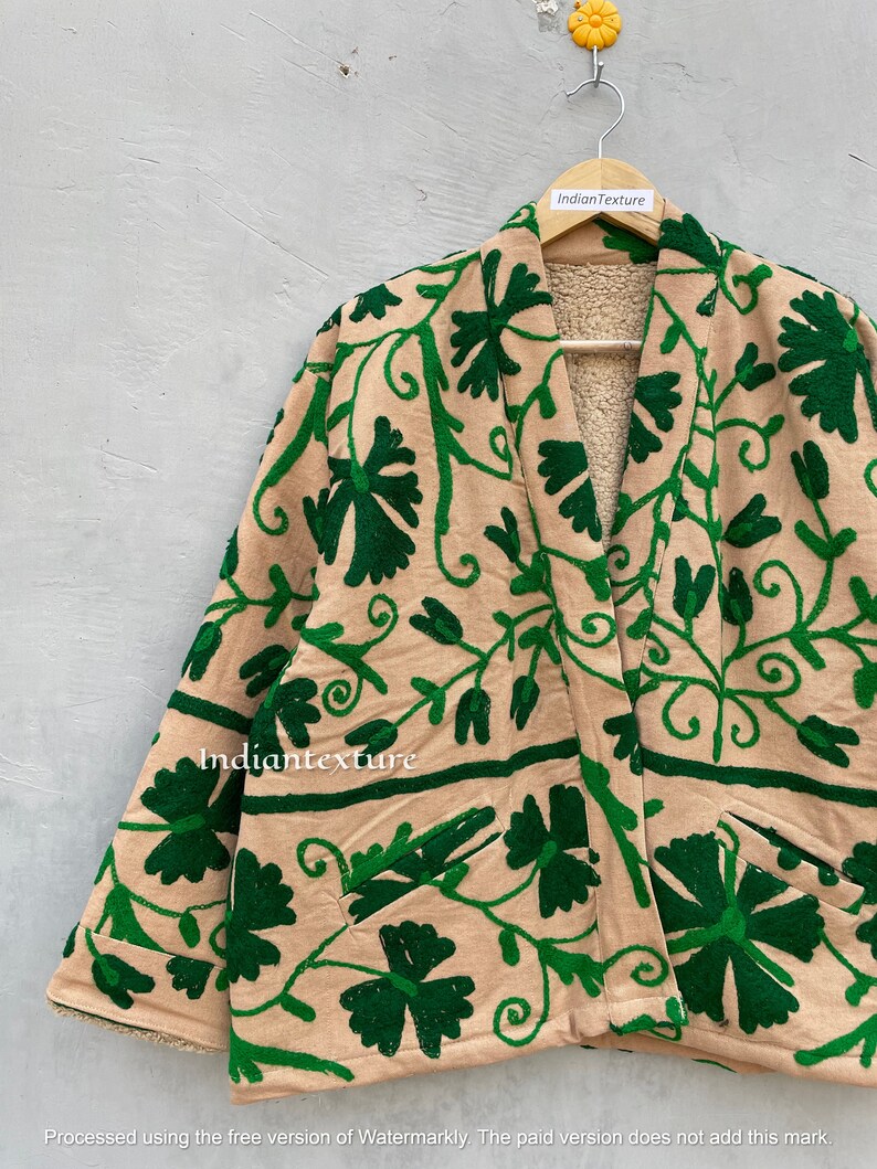 Op de afbeelding: Een beige jas met een groen geborduurd bloemenpatroon. De jas heeft een shawlkraag en lange mouwen. De jas is gevoerd met een zacht, bruin fleece.