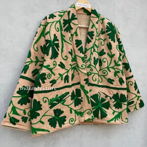 Op de afbeelding: Een beige jas met een groen geborduurd bloemenpatroon. De jas heeft een shawlkraag en lange mouwen. De jas is gevoerd met een zacht, pluche materiaal.