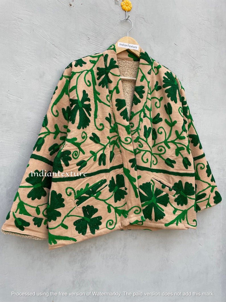 Op de afbeelding: Een beige jas met een groen geborduurd bloemenpatroon. De jas heeft een shawlkraag en lange mouwen. De jas is gevoerd met een zacht, pluche materiaal.