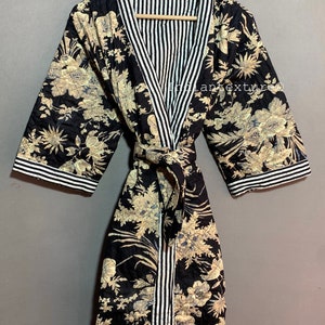 May include: A black quilted robe with a floral pattern in beige and blue. The robe features a striped trim in black and white along the collar, cuffs, and belt. The robe is displayed on a wooden hanger.
