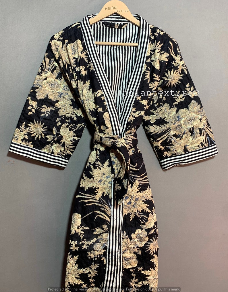 May include: A black quilted robe with a floral pattern in beige and blue. The robe features a striped trim in black and white along the collar, cuffs, and belt. The robe is displayed on a wooden hanger.