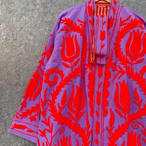 Puede incluir: Una larga t&uacute;nica morada con dise&ntilde;os florales bordados en rojo. La t&uacute;nica tiene un forro a rayas rojo y blanco visible en el cuello.